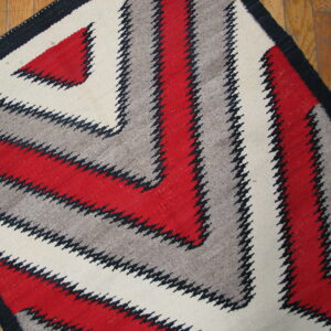 A low-pile rug with red, gray, cream, and black serrated zig-zags rests on a wood plank floor.