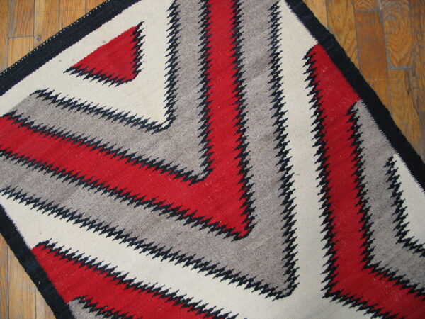 A low-pile rug with red, gray, cream, and black serrated zig-zags rests on a wood plank floor.