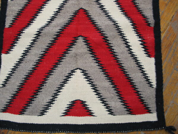 Flatweave rug with serrated red, gray, and cream zigzags bordered in black, resting on wood planks.
