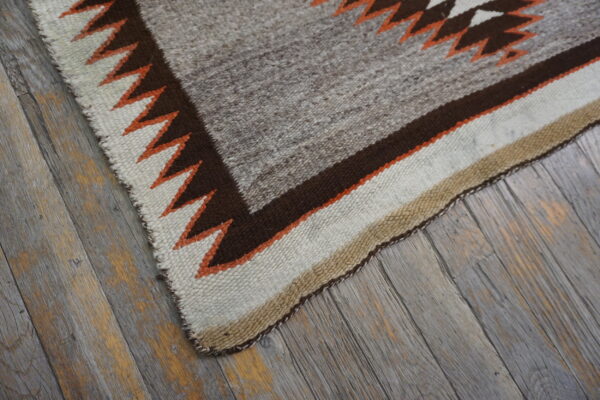 Flatwoven gray rug with brown, cream, and orange geometric border lying on distressed wood flooring.