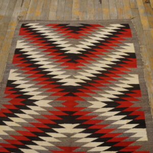 A flatweave rug featuring red, black, white, and gray geometric diamond patterns on worn plank flooring.