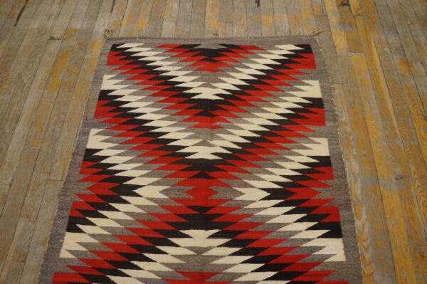 Bold tribal geometric pattern antique native american flat weave navajo rug 4 A flatweave rug featuring red, black, white, and gray geometric diamond patterns on worn plank flooring.