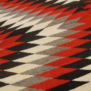 A flat-woven rug displays red, black, cream, and gray geometric diamond shapes with a solid border.