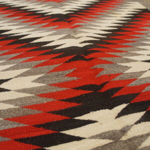 Low-pile rug with red, black, grey, and cream serrated diamond patterns against a wood floor.
