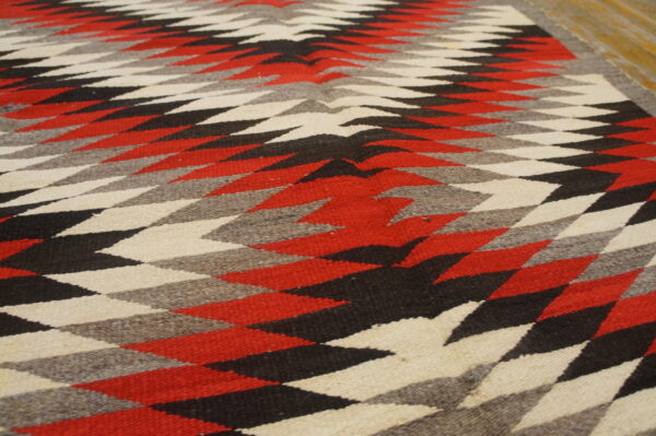 Bold tribal geometric pattern antique native american flat weave navajo rug 6 Low-pile rug with red, black, grey, and cream serrated diamond patterns against a wood floor.