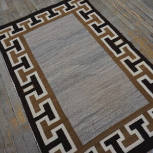 Flatweave rug with a mottled gray center and a dark geometric Greek key border on rustic wood.