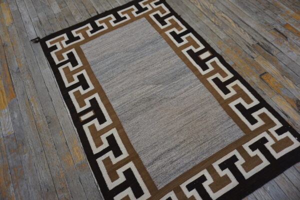 Flatweave rug with a mottled gray center and a dark geometric greek key border on rustic wood.