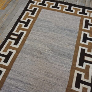 Runner rug with a gray central field and geometric border in dark brown and tan on distressed wood floor.