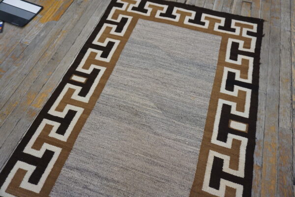 Runner rug with a gray central field and geometric border in dark brown and tan on distressed wood floor.