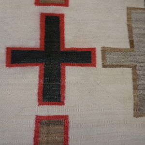 Close-up of a cream woven textile featuring a geometric black and red cross design next to weathered wood.