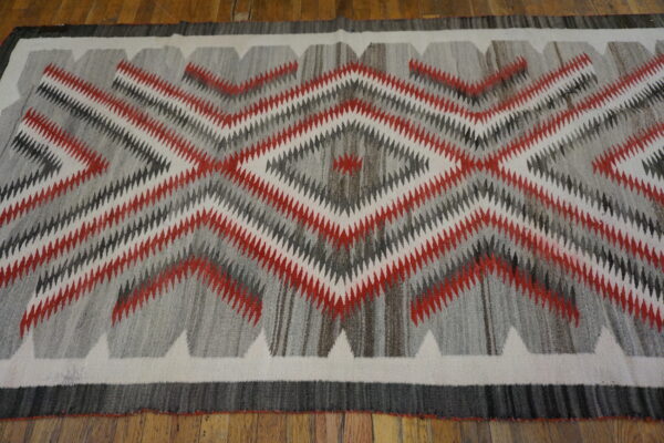 Red, white, and charcoal rug featuring serrated diamond patterns lies flat on wood planks.