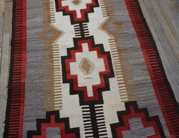 Flat-woven rug with red, black, white, and gray geometric designs on distressed wooden flooring.