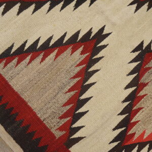 Flatweave rug with serrated red, brown, and cream geometric patterns rests on light wood planks.