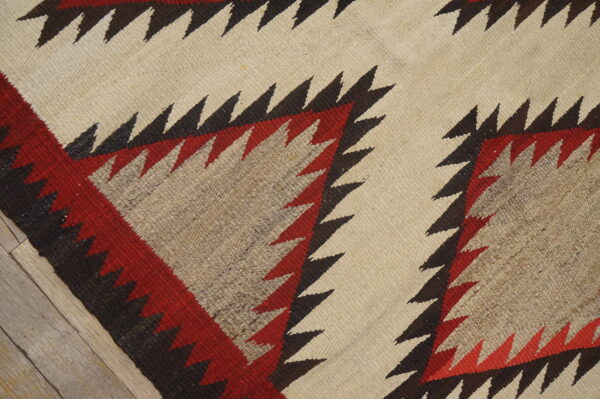 Flatweave rug with serrated red, brown, and cream geometric patterns rests on light wood planks.