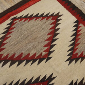 Cream, red, and brown flatweave rug with serrated diamond pattern resting on rustic wood planks.