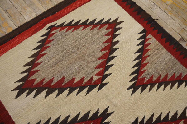 Cream, red, and brown flatweave rug with serrated diamond pattern resting on rustic wood planks.
