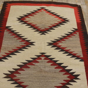 Cream, taupe, red, and dark brown flatweave rug with serrated diamond patterns on worn wood floor.