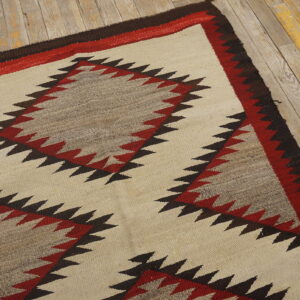 Cream flatweave rug with jagged red, brown, and gray geometric designs on worn wood plank flooring.