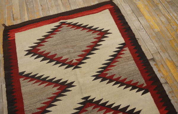 Cream flatweave rug with jagged red, brown, and gray geometric designs on worn wood plank flooring.