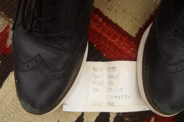 Black leather shoes and a white information tag rest on a patterned red, tan, and brown flatwoven rug.