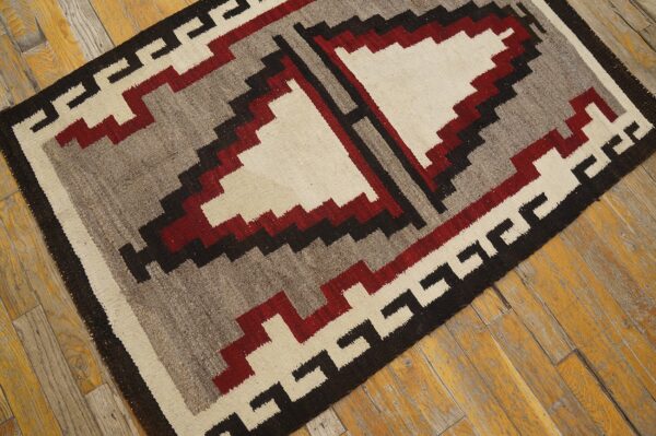 Geometric flatweave rug featuring stepped patterns in cream, red, gray, and black on distressed wood flooring.
