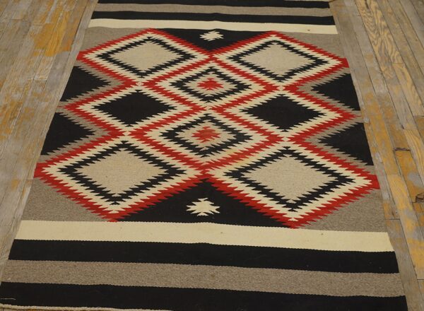 Black, red, and beige flat-weave rug with serrated diamonds lies on weathered wooden plank flooring.