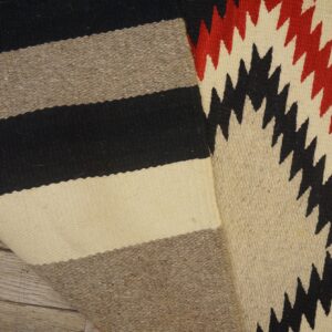Flatweave rug folded, showing neutral stripes and a red and black geometric zigzag pattern on wood.