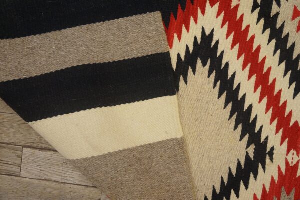 Flatweave rug folded, showing neutral stripes and a red and black geometric zigzag pattern on wood.
