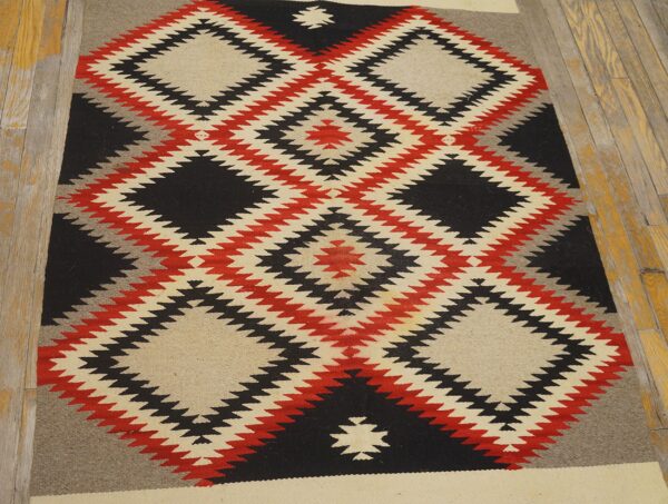Flatweave rug in black, red, gray, and cream geometric diamond patterns rests on wood flooring.