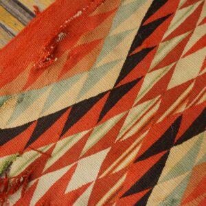 Geometric flatweave rug featuring red, cream, green, and black diagonal zigzags resting on rustic wood flooring.