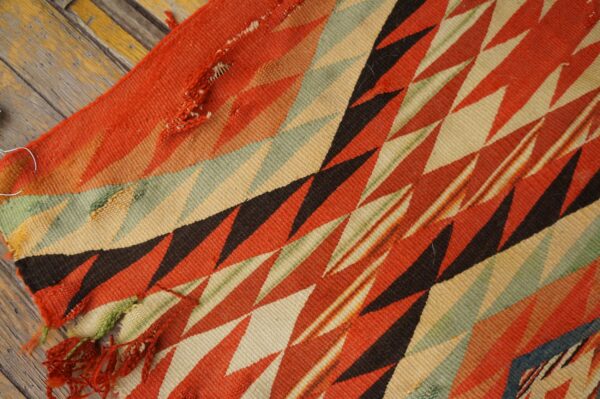 Geometric flatweave rug featuring red, cream, green, and black diagonal zigzags resting on rustic wood flooring.