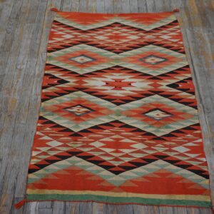 Low-pile rug with orange, black, and cream geometric pattern sits on worn gray wood flooring.