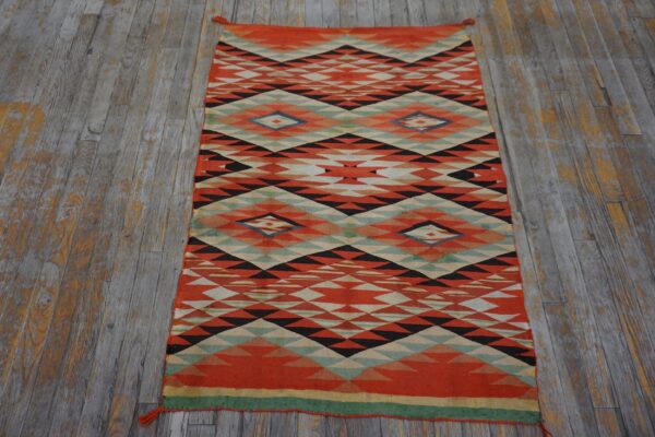 Low-pile rug with orange, black, and cream geometric pattern sits on worn gray wood flooring.
