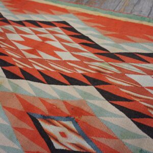 Geometric flat weave rug featuring red, black, and teal zigzag patterns lying on weathered gray planks.