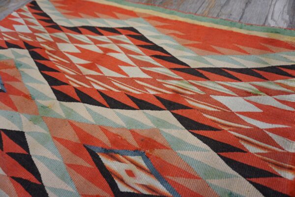 Geometric flat weave rug featuring red, black, and teal zigzag patterns lying on weathered gray planks.