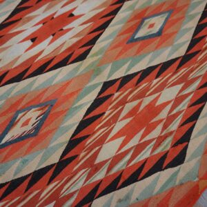 Flat woven rug with sharp geometric patterns in rust, black, cream, and teal on distressed wooden planks.