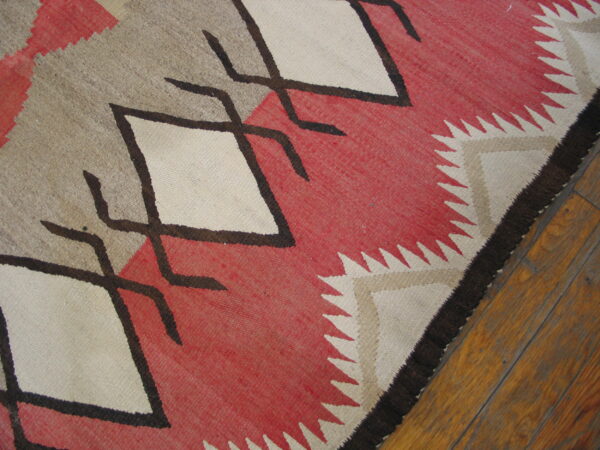 A flatweave rug features bold red, tan, and white diamonds outlined in dark brown on wood planks.