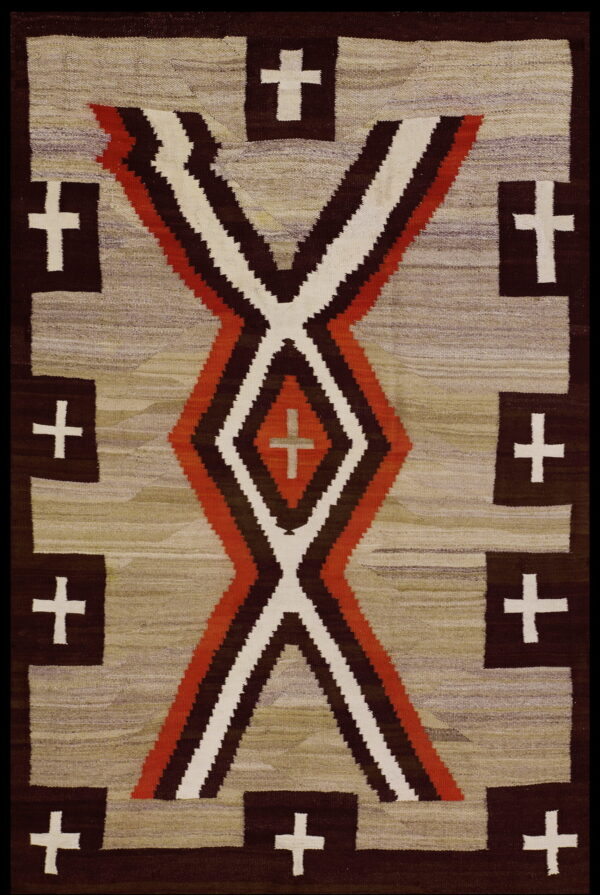 Flat woven rug with rust, brown, and cream hourglass shapes surrounded by crosses on a mottled tan field.