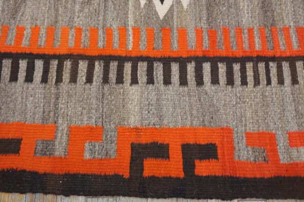 Close-up of flatweave rug featuring interlocking orange and black geometric designs on a mixed gray background.