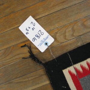 A flatweave rug corner featuring black, red, cream, and gray jagged patterns rests on distressed wooden flooring.