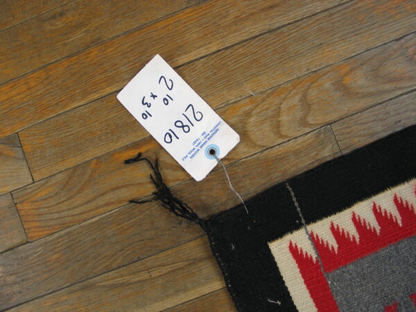 A flatweave rug corner featuring black, red, cream, and gray jagged patterns rests on distressed wooden flooring.