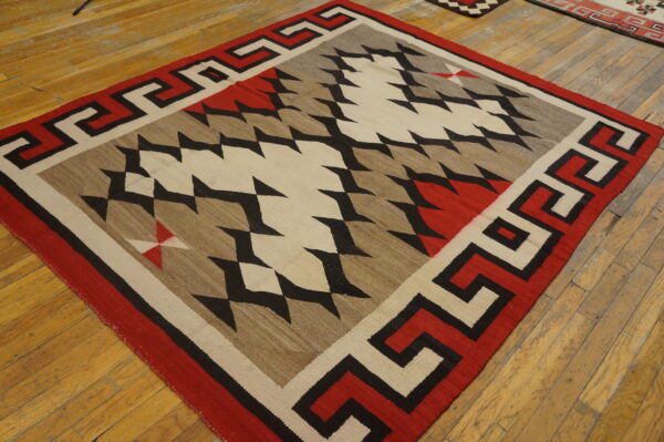 Red, black, beige, and brown geometric flatweave rug with a meander border on light wood flooring.