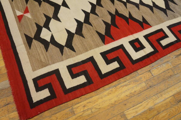Geometric flatweave rug featuring red, black, tan, and cream serrated patterns on distressed wooden floorboards.