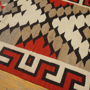A flatweave rug featuring red, black, cream, and tan geometric diamonds and a Greek key border on wood floor.