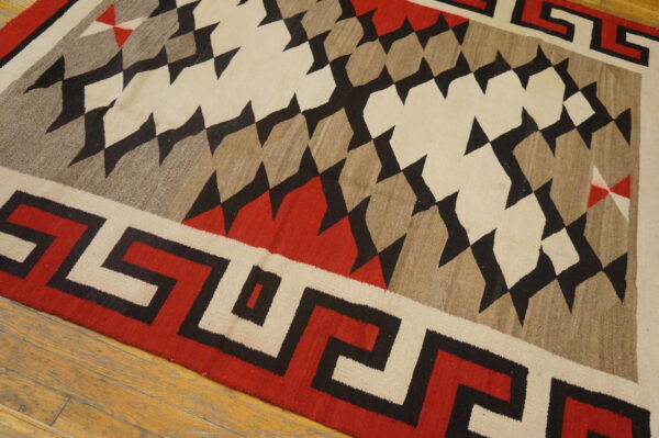 A flatweave rug featuring red, black, cream, and tan geometric diamonds and a greek key border on wood floor.