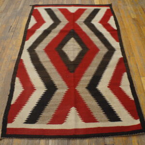 Flatweave rug with serrated red, black, cream, and gray geometric patterns rests on worn wood flooring.