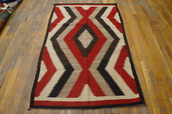 Flatweave rug with serrated red, black, cream, and gray geometric patterns rests on worn wood flooring.