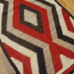 Flatweave rug in red, black, beige, and tan with serrated diamond designs lying on light wooden flooring.