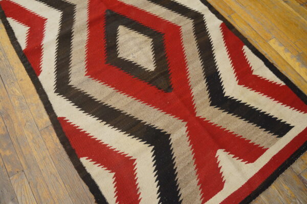 Flatweave rug in red, black, beige, and tan with serrated diamond designs lying on light wooden flooring.