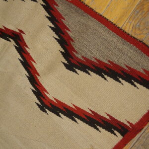 Beige flatweave rug with serrated red and black geometric designs on rustic wooden floors.